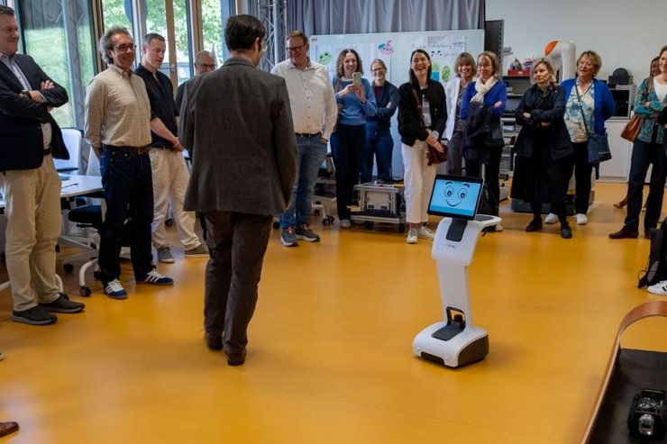 Prof. Dr. Manuel Giuliani demonstriert einer Delegation des Bayerischen Wissenschaftsministeriums im Care and Mobile Robots Lab der Hochschule Kempten einen mobilen Kommunikationsroboter mit Bildschirmgesicht. Die Gäste stehen im Halbkreis um den Roboter und verfolgen die Vorführung. Im Vordergrund ist ein weiterer kleiner Roboter auf einer Teststrecke zu sehen. Die Aufnahme zeigt den Besuch der für Hochschulen für angewandte Wissenschaften zuständigen Abteilung des Ministeriums und gibt Einblicke in die Forschung zu Assistenzrobotik und mobilen Robotersystemen.