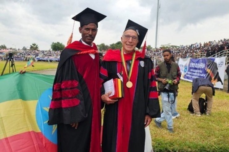 Prof. Dr.-Ing. Dr. h. c. mult. Johannes Steinbrunn steht gemeinsam mit einem Vertreter der Hawassa University in akademischer Festkleidung auf dem Veranstaltungsgelände der Universität. Steinbrunn trägt die Insignien seiner Ehrendoktorwürde und hält die Verleihungsurkunde in den Händen. Im Hintergrund sind Gäste und Zuschauer der akademischen Feier zu sehen. Die Hawassa University verlieh ihm den Ehrendoktortitel (Doctor of Science honoris causa) für seine langjährigen Verdienste um Ingenieurwissenschaften, Hochschulentwicklung und die internationale wissenschaftliche Zusammenarbeit.