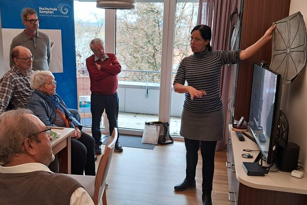 Mehrere ältere Personen hören einer Frau beim Reden zu. Diese hälte eine quadratische Matte in die Höhe.